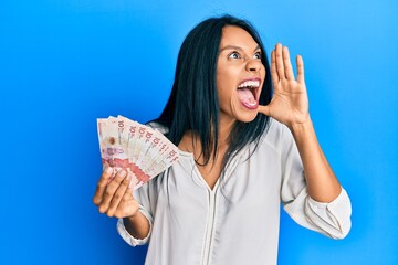 Young african american woman holding 10 colombian pesos banknotes shouting and screaming loud to side with hand on mouth. communication concept.