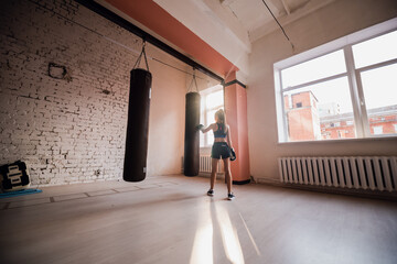A pretty kickboxer girl is preparing for competitions in the boxing hall, practicing the technique of punches on a punching bag