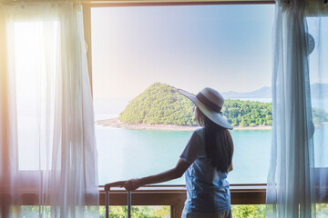 asian tourist woman standing nearly window looking and relax with beautiful view with her luggage in hotel bedroom after check-in. Happy women with holiday summer travel vacation concept
