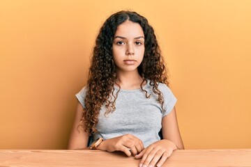 Teenager hispanic girl wearing casual clothes sitting on the table relaxed with serious expression...