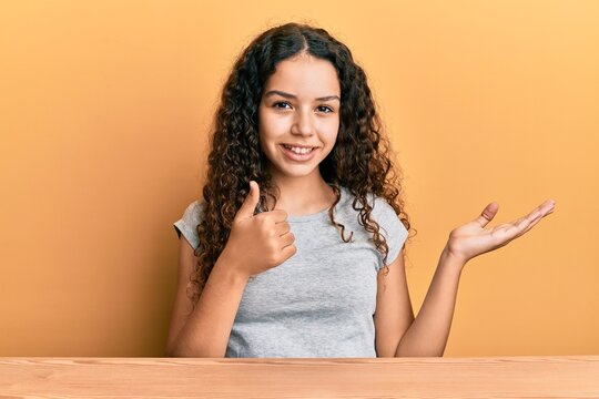 Teenager hispanic girl wearing casual clothes sitting on the table showing palm hand and doing ok gesture with thumbs up, smiling happy and cheerful