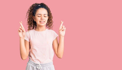 Beautiful kid girl with curly hair wearing casual clothes gesturing finger crossed smiling with hope and eyes closed. luck and superstitious concept.