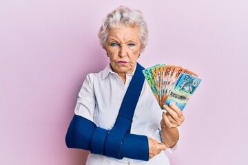 Senior grey-haired woman wearing arm on sling holding australia dollars banknotes depressed and worry for distress, crying angry and afraid. sad expression.