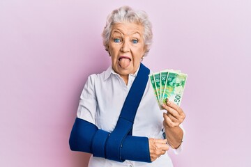 Senior grey-haired woman wearing arm on sling holding israel shekels banknotes sticking tongue out...