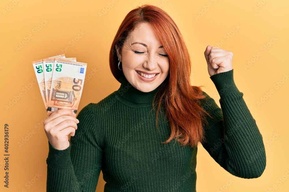 Poster Beautiful redhead woman holding bunch of 50 euro banknotes screaming proud, celebrating victory and success very excited with raised arm