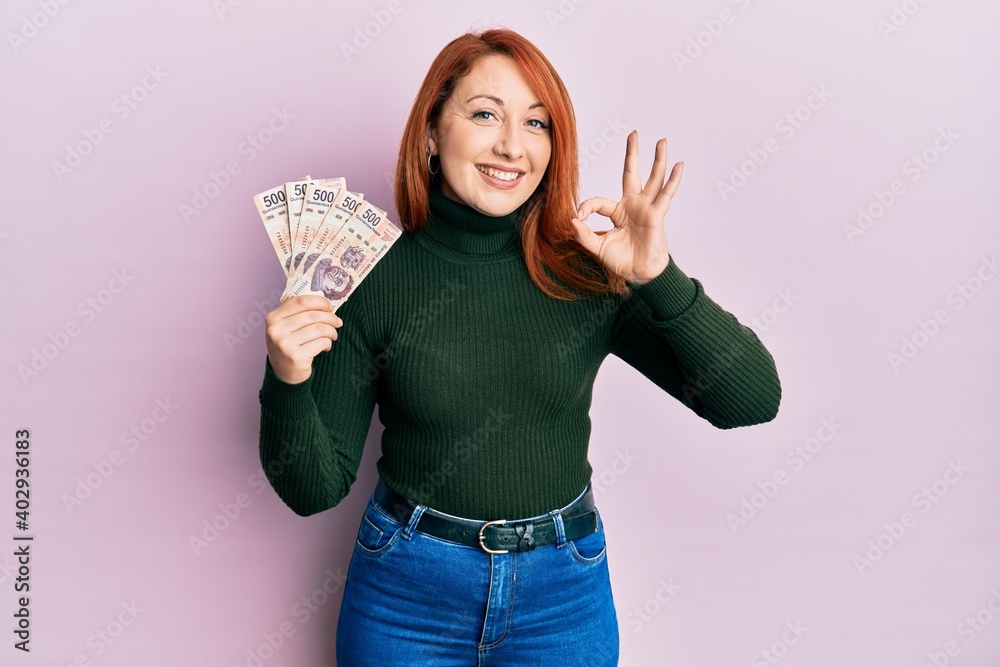 Canvas Prints Beautiful redhead woman holding 500 mexican pesos banknotes doing ok sign with fingers, smiling friendly gesturing excellent symbol