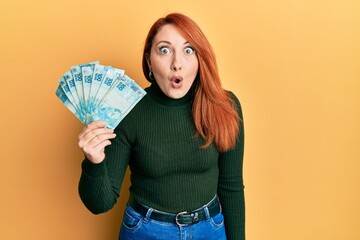 Beautiful redhead woman holding 100 brazilian real banknotes scared and amazed with open mouth for...