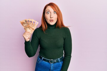 Beautiful redhead woman holding south african 20 rand banknotes scared and amazed with open mouth for surprise, disbelief face