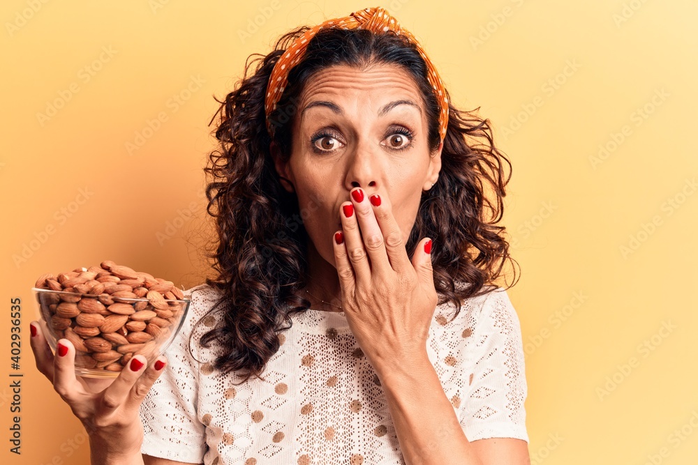 Wall mural middle age beautiful woman holding bowl with almonds covering mouth with hand, shocked and afraid fo