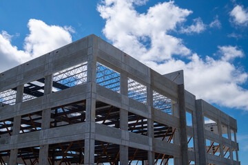A large multiple storey precast grey concrete building with steel beams against a blue sky. The industrial structure is the corner of a skyscraper building with prefabricated engineering formwork. - obrazy, fototapety, plakaty