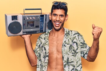 Young latin man wearing summer shirt holding boombox screaming proud, celebrating victory and success very excited with raised arms