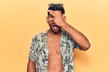 Young latin man wearing summer shirt and sunglasses peeking in shock covering face and eyes with hand, looking through fingers afraid