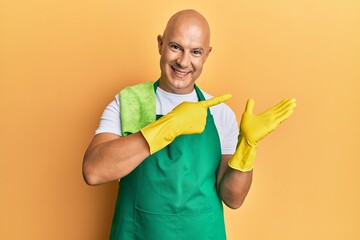 Middle age bald man wearing apron holding cleaning gloves amazed and smiling to the camera while presenting with hand and pointing with finger.