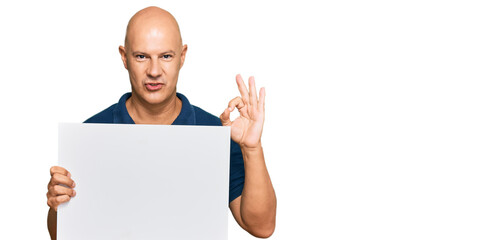 Middle age bald man holding blank empty banner doing ok sign with fingers, smiling friendly gesturing excellent symbol