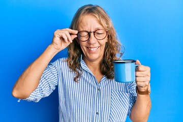Middle age blonde woman wearing glasses and drinking a cup of coffee winking looking at the camera...