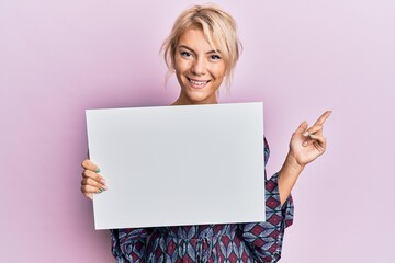 Naklejka premium Young blonde girl holding blank empty banner smiling happy pointing with hand and finger to the side