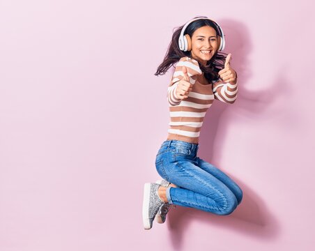 Young beautiful hispanic woman smiling happy. Jumping with smile on face listening to music using headphones doing ok sign with thums up over pink background