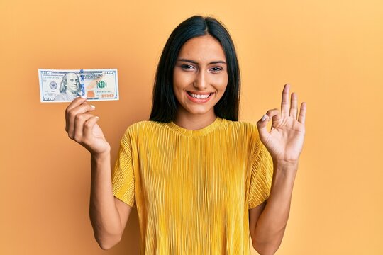 Young brunette woman holding 100 dollars banknote doing ok sign with fingers, smiling friendly gesturing excellent symbol