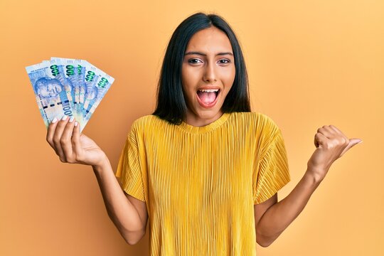 Young brunette woman holding south african 100 rand banknotes pointing thumb up to the side smiling happy with open mouth