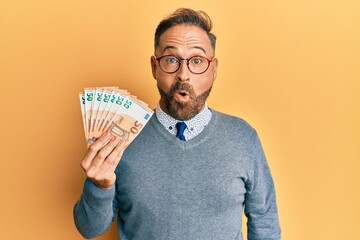 Handsome middle age man holding bunch of 50 euro banknotes scared and amazed with open mouth for surprise, disbelief face - obrazy, fototapety, plakaty