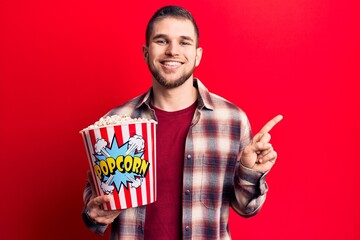 Young handsome man eating popcorn smiling happy pointing with hand and finger to the side
