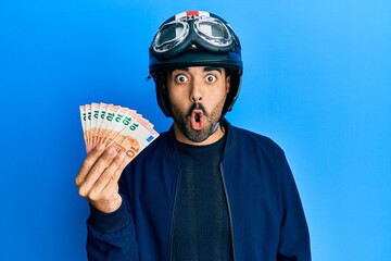 Young hispanic man wearing motorcycle helmet holding 10 euros scared and amazed with open mouth for...