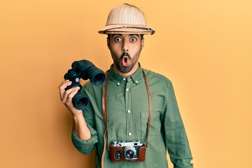 Young hispanic man wearing explorer hat looking through binoculars scared and amazed with open mouth for surprise, disbelief face - obrazy, fototapety, plakaty