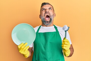 Middle age handsome man wearing apron holding scourer washing dishes angry and mad screaming frustrated and furious, shouting with anger looking up.