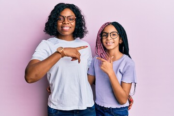 Beautiful african american mother and daughter wearing casual clothes and glasses cheerful with a smile on face pointing with hand and finger up to the side with happy and natural expression