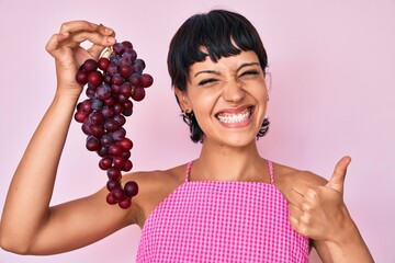 Beautiful brunettte woman holding branch of fresh grapes smiling happy and positive, thumb up doing excellent and approval sign