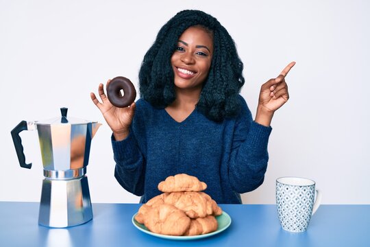 Beautiful african woman eating breakfast holding cholate donut smiling happy pointing with hand and finger to the side