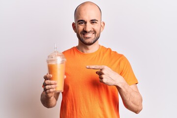 Young handsome bald man drinking glass of healthy orange juice smiling happy pointing with hand and finger