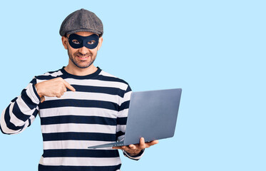 Young handsome man wearing burglar mask using laptop pointing finger to one self smiling happy and proud