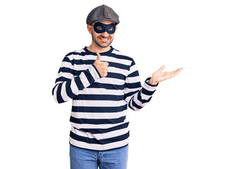 Young handsome man wearing burglar mask showing palm hand and doing ok gesture with thumbs up, smiling happy and cheerful