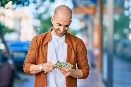 Young hispanic bald man smiling happy counting usa dollars at the city.