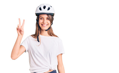 Young beautiful girl wearing bike helmet smiling with happy face winking at the camera doing victory sign. number two.