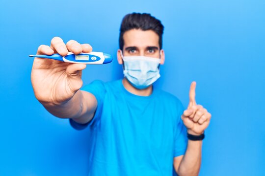 Young handsome man with beard wearing medical mask holding thermometer smiling with an idea or question pointing finger with happy face, number one
