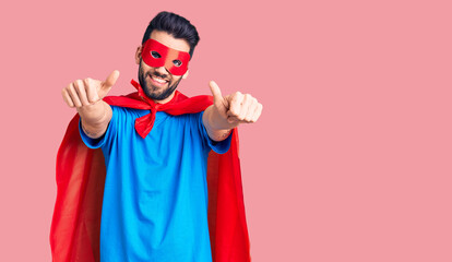 Young handsome man with beard wearing super hero costume approving doing positive gesture with hand, thumbs up smiling and happy for success. winner gesture.