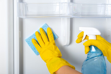 A woman's hands in a yellow rubber protective glove and a blue sponge washes, cleans refrigerator shelves. Cleaning service, housewife, routine housework. Spray for windows and glass surfaces cleaner