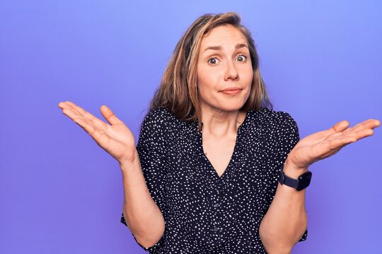 Young beautiful blonde woman standing over purple isolated background clueless and confused with open arms, no idea and doubtful face.
