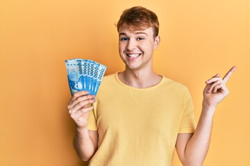Young caucasian man holding mexican pesos smiling happy pointing with hand and finger to the side