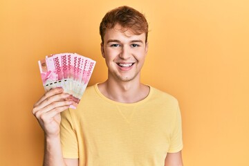 Young caucasian man holding 100000 indonesian rupiah looking positive and happy standing and smiling with a confident smile showing teeth