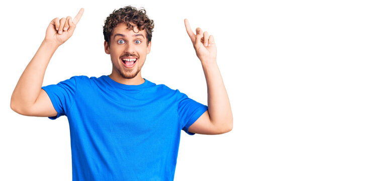 Young handsome man with curly hair wearing casual clothes smiling amazed and surprised and pointing up with fingers and raised arms.
