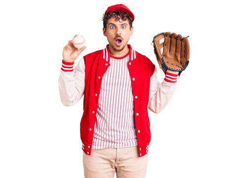Young handsome man with curly hair wearing baseball uniform holding golve and ball scared and amazed with open mouth for surprise, disbelief face