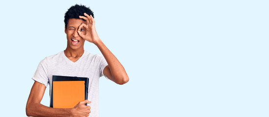 Young african american man holding book smiling happy doing ok sign with hand on eye looking through fingers