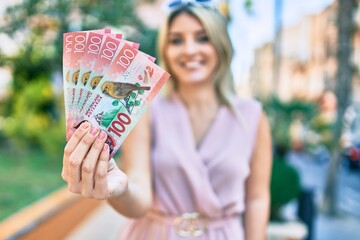 Young blonde woman smiling happy holding new zealand 100 dollars banknotes at the city.