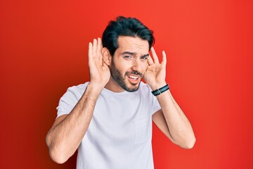 Fototapeta premium Young hispanic man wearing casual white tshirt trying to hear both hands on ear gesture, curious for gossip. hearing problem, deaf
