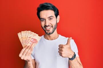 Young hispanic man holding mexican pesos smiling happy and positive, thumb up doing excellent and...