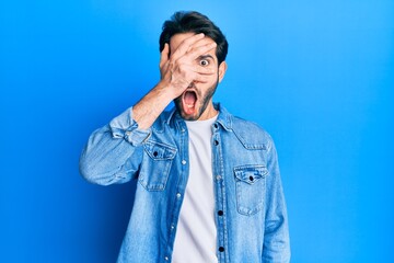Young hispanic man wearing casual clothes peeking in shock covering face and eyes with hand,...