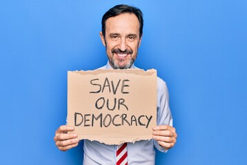 Middle age business man wearing usa tie holding banner with save our democracy message looking positive and happy standing and smiling with a confident smile showing teeth